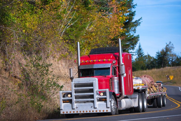 Powerful heavy huge bright red professional classical big rig semi truck with a thick protective grille and high tailpipes and step-down trailer where the straps tied several pallets load at the turn of autumn winding road.